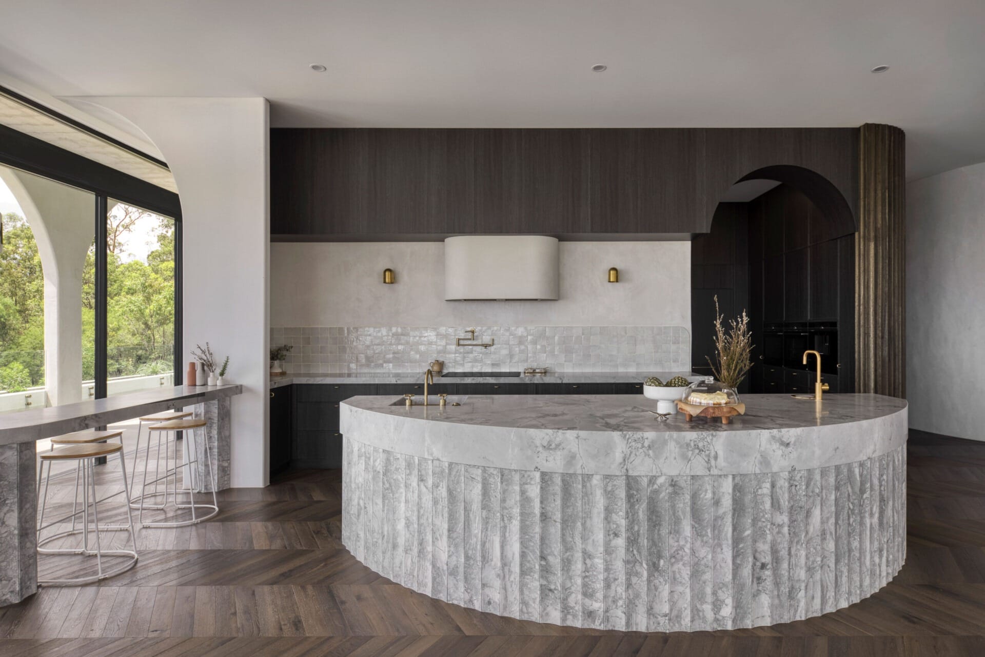 Luxury kitchen featuring dark herringbone timber flooring, curved marble island, and deep-toned cabinetry.