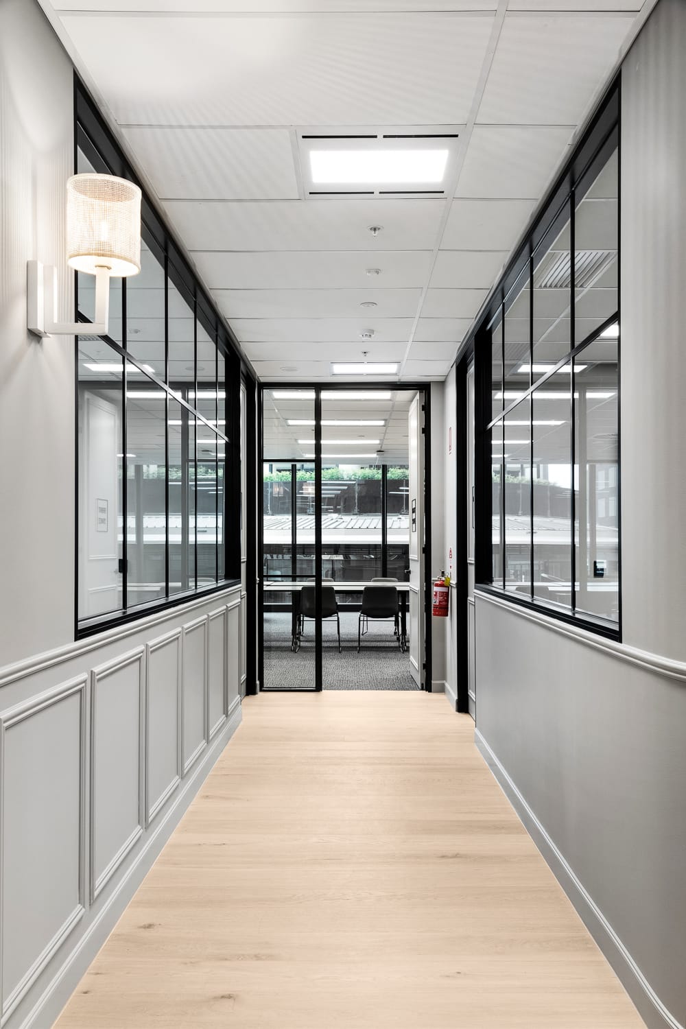 Light engineered timber flooring in a commercial hallway with grey wall panelling and black-framed glass office partitions.