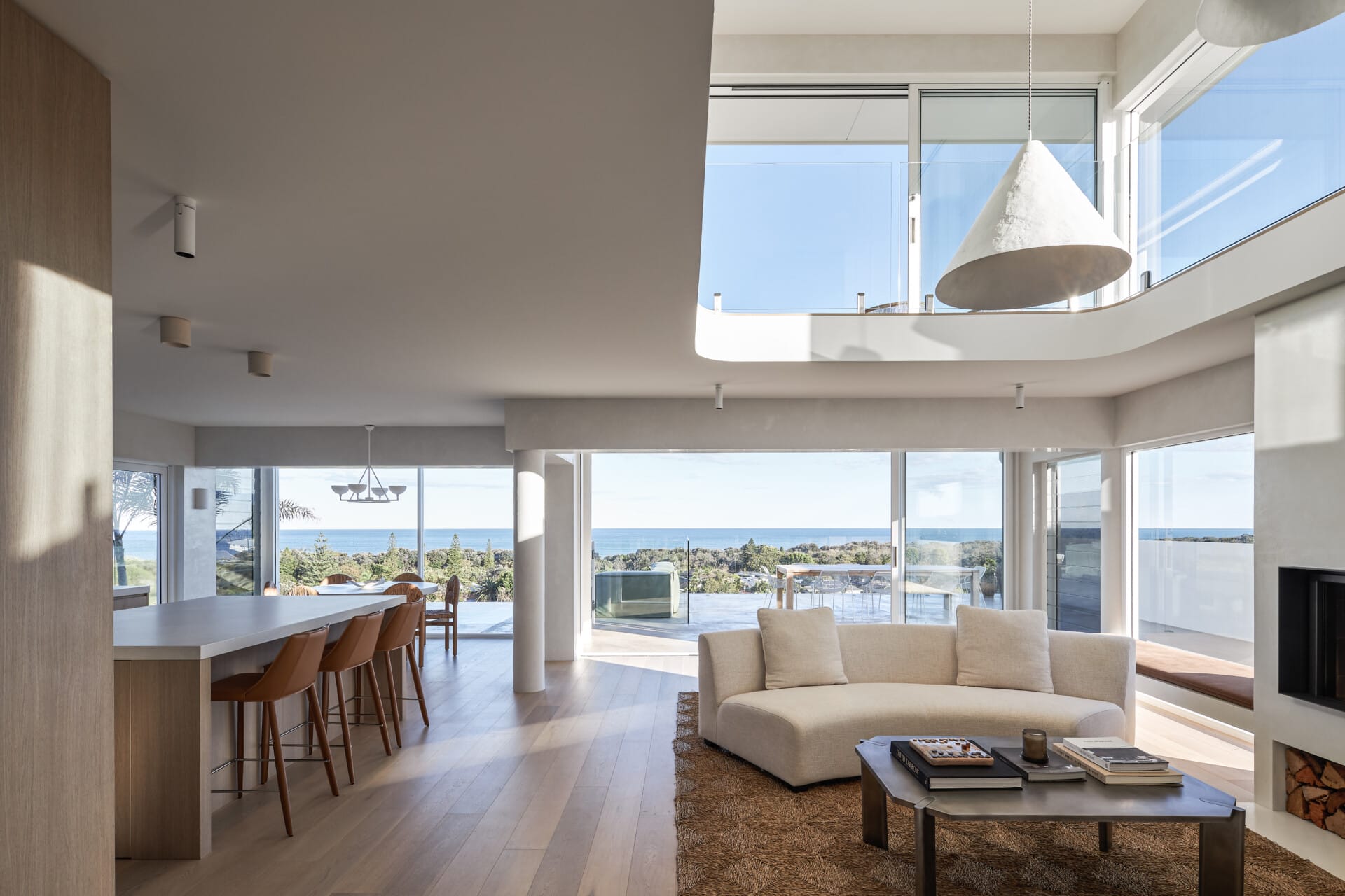 Open-plan coastal living space with wide plank timber flooring, curved sofa, large windows and ocean views.