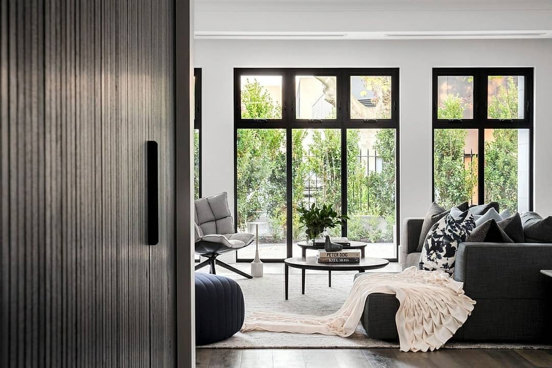 Modern living room with dark timber flooring, black-framed windows, neutral furnishings and soft natural light overlooking a garden.
