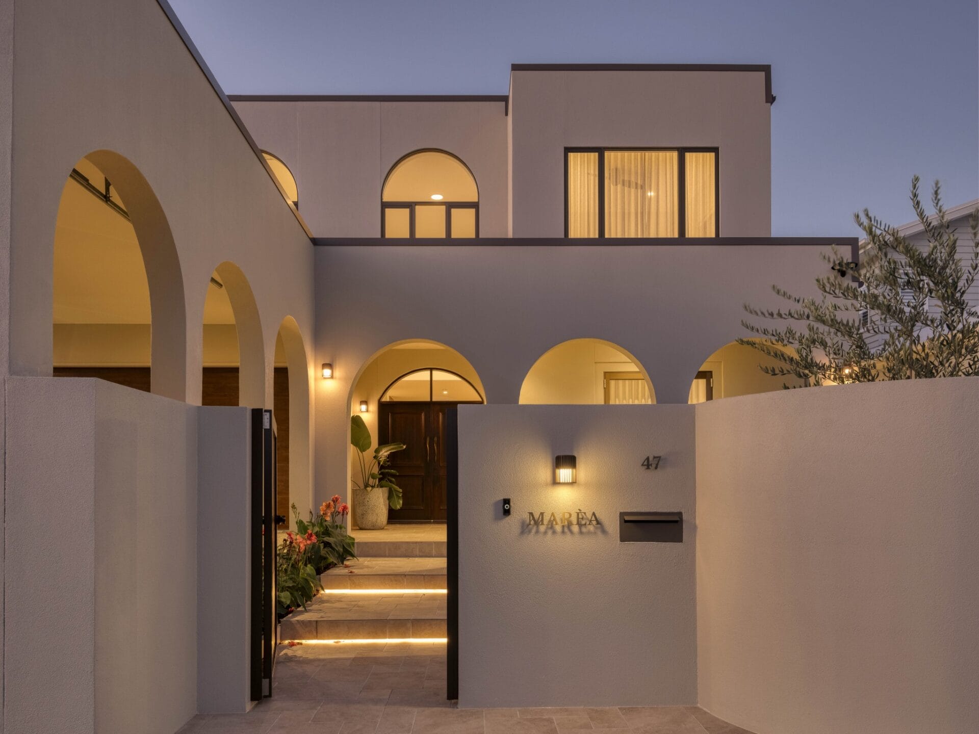Entry courtyard at MARÈA with layered arches, landscaped steps and brass signage at dusk