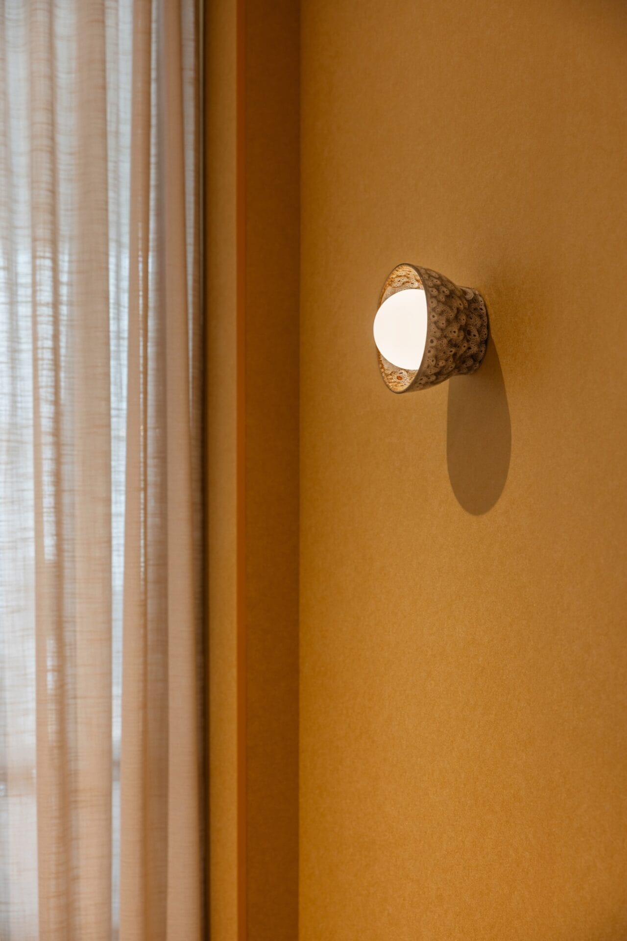 Wall sconce mounted on warm textured render beside a sheer curtain