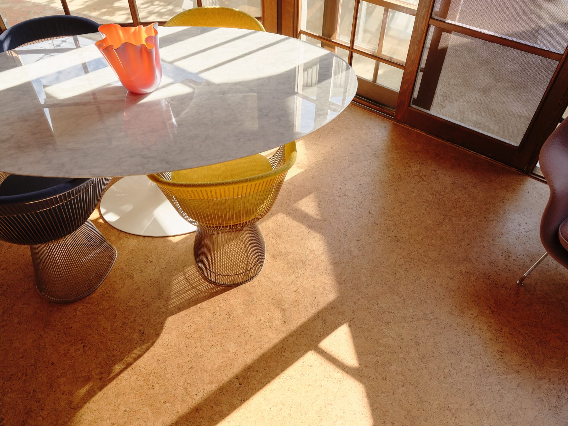 Oval marble dining table with an orange glass vase and black and yellow Platner chairs, Porto cork flooring catching afternoon light.