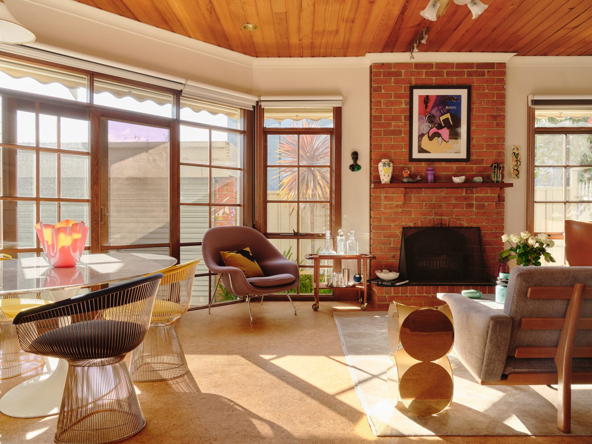 Sunroom dining nook with black wire-frame Platner chairs and a woven rattan armchair, French doors opening to a palm-lined garden, brick fireplace with framed artwork, Porto cork flooring throughout.