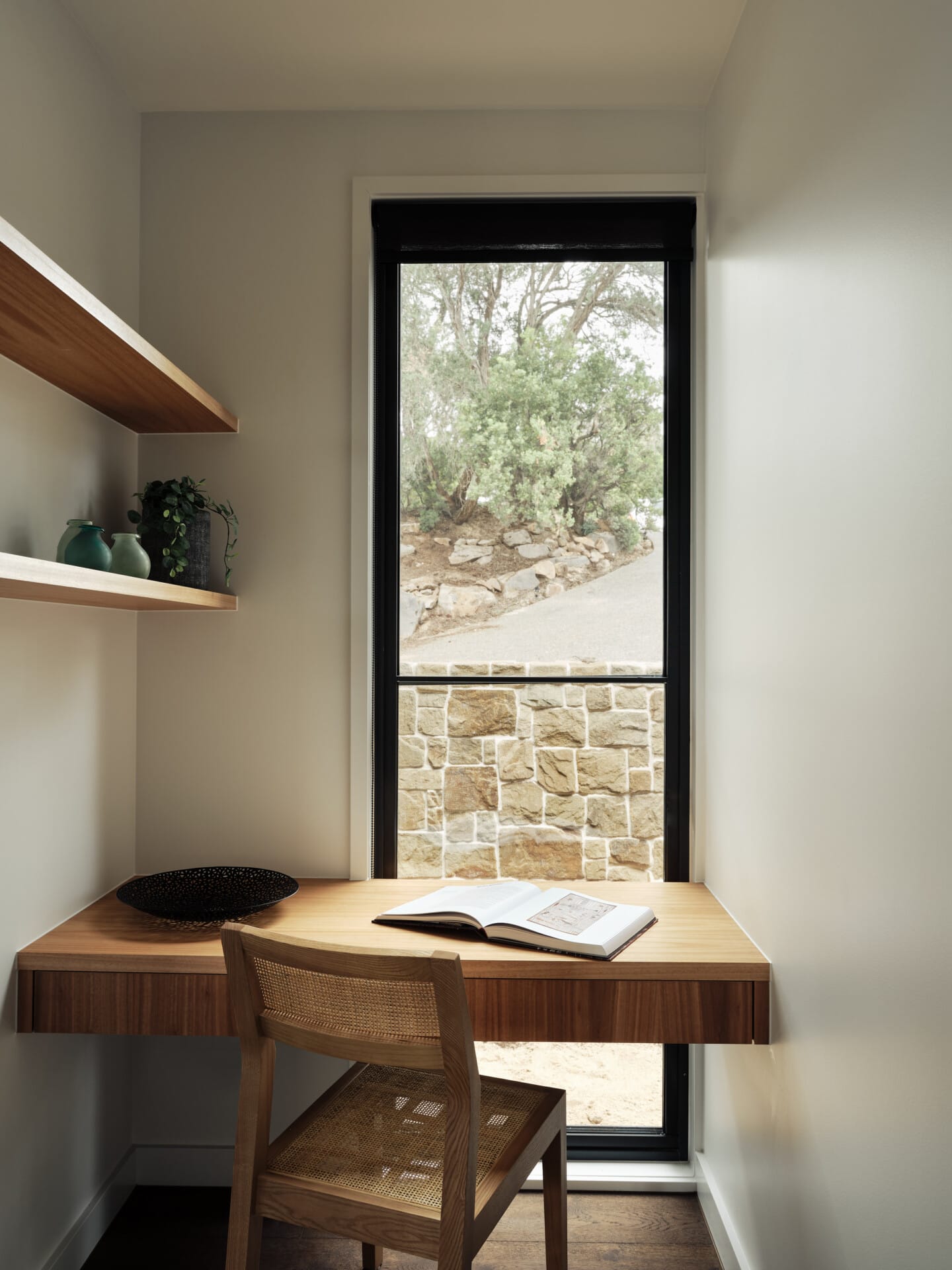 Built-in timber study nook with a floating desk, cane chair, and floor-to-ceiling window framing a view of the stone garden wall.