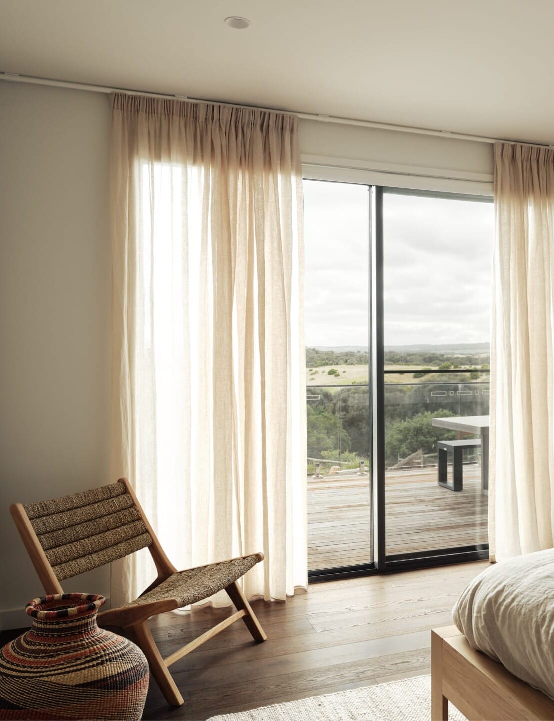 Bedroom with sheer curtains framing a sliding door to a timber deck, Le Cru 190mm timber flooring, a woven armchair and basket in the corner.