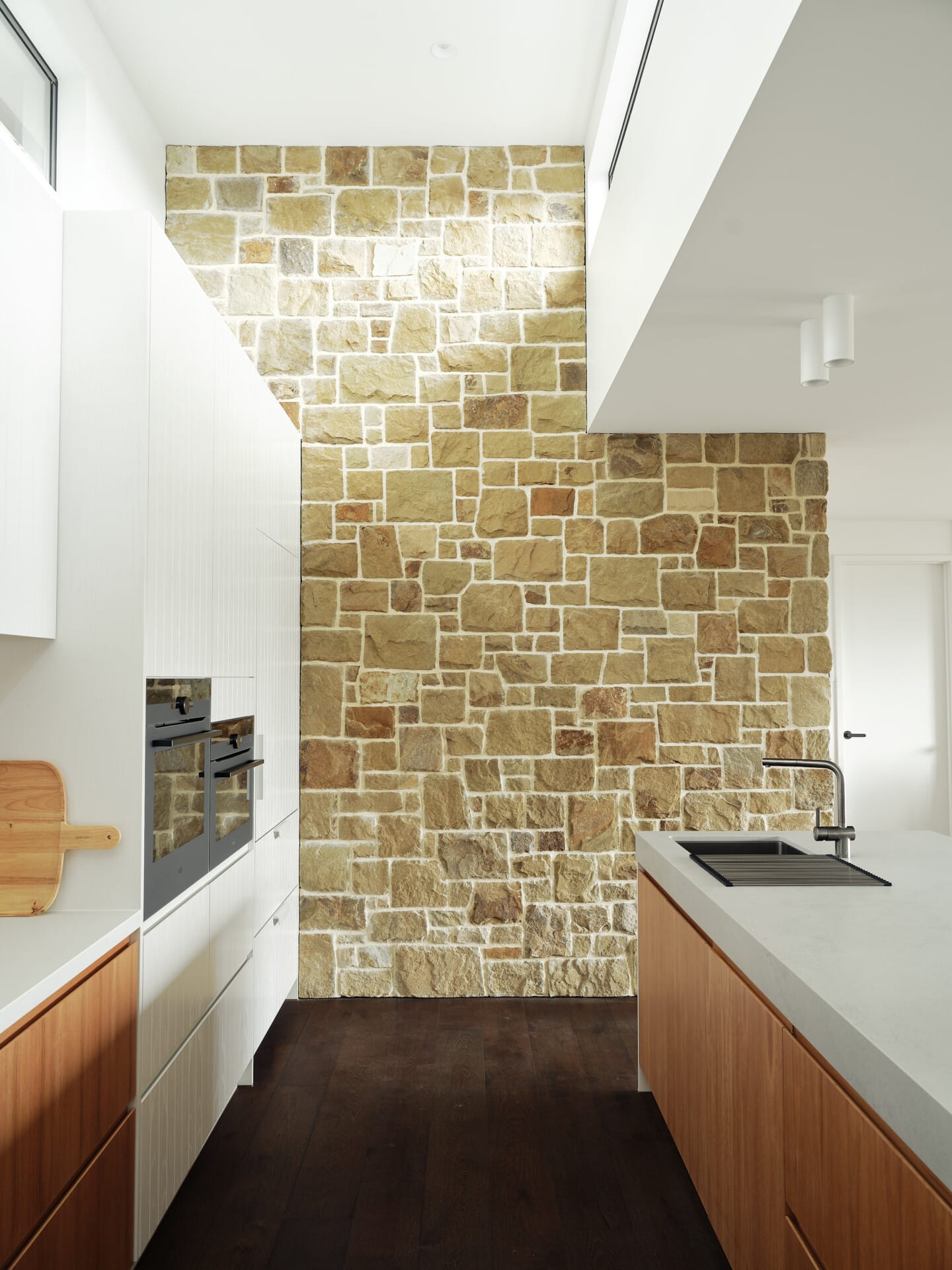 Galley kitchen with a full-height stone feature wall, timber and white cabinetry either side, Le Cru 190mm timber flooring underfoot.