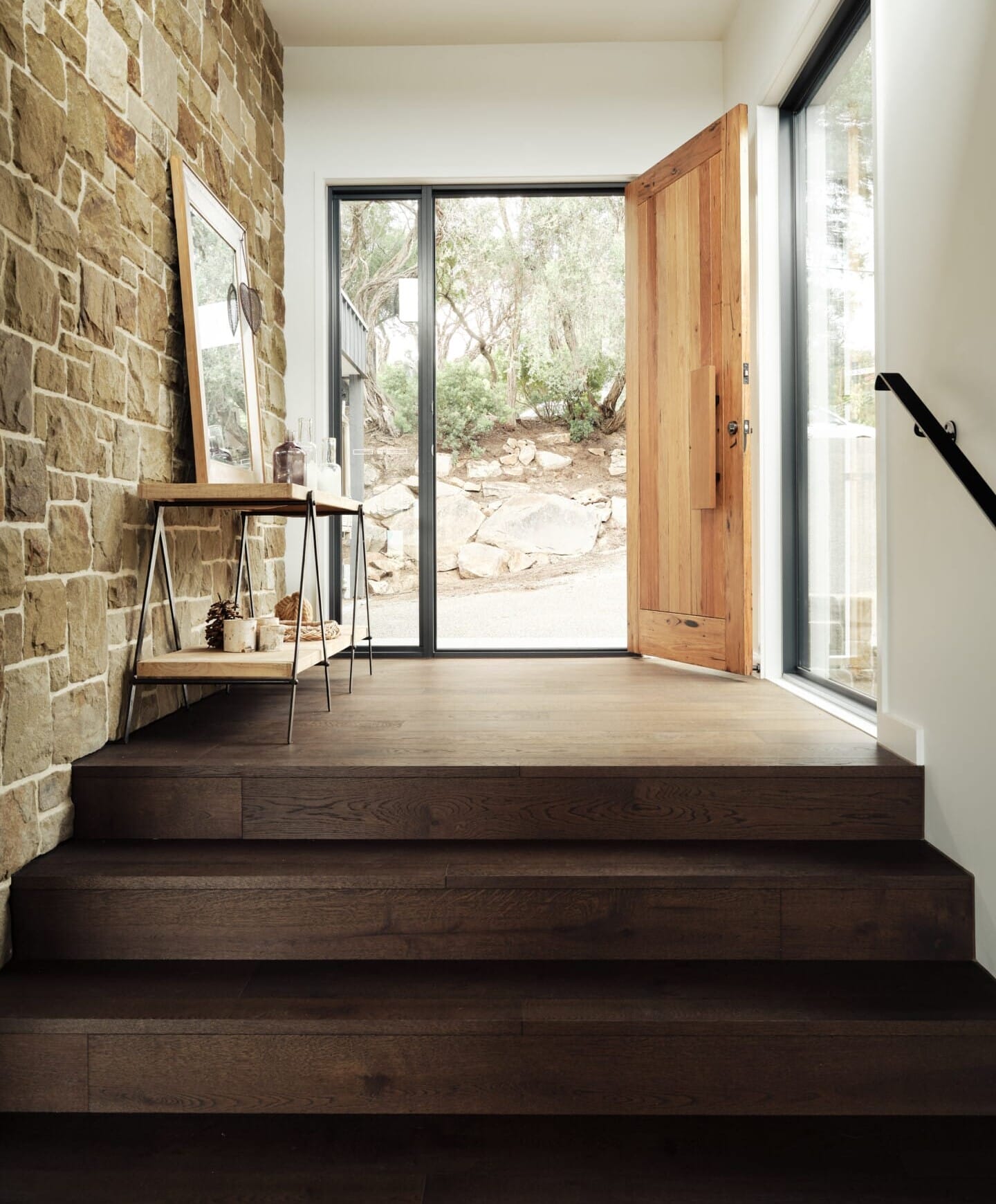 Front entrance at St Andrews Residence with a stone feature wall, an oak door open to a garden view, dark Le Cru 190mm timber flooring on the steps down to a staircase with a black handrail.