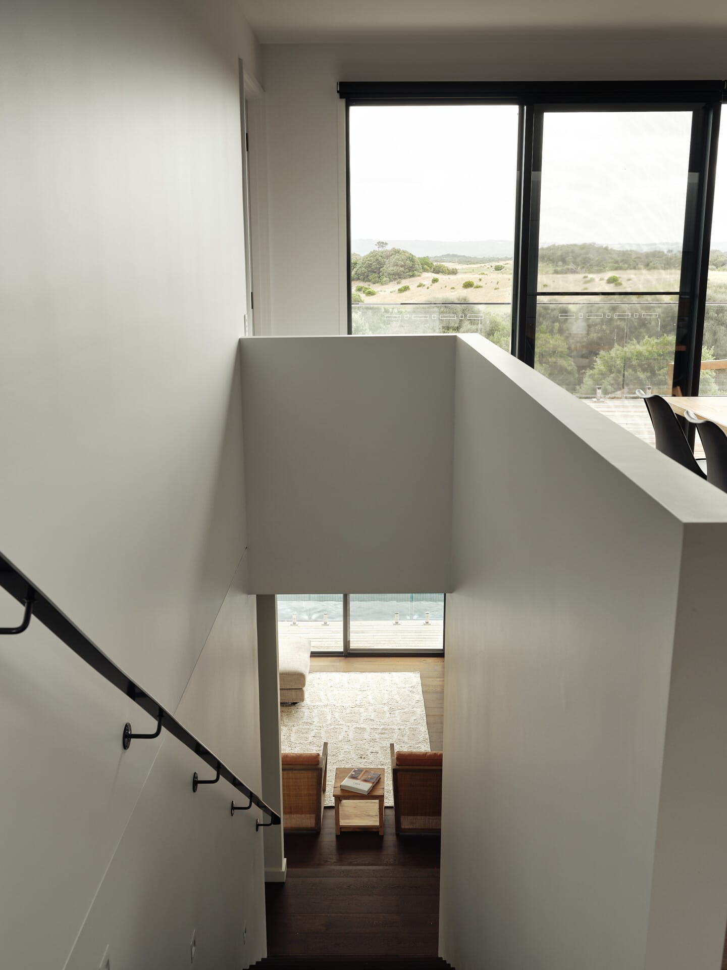 View down an internal staircase past a white balustrade to a lounge with rattan armchairs, Le Cru 190mm timber flooring underfoot, golf course views through the window above.