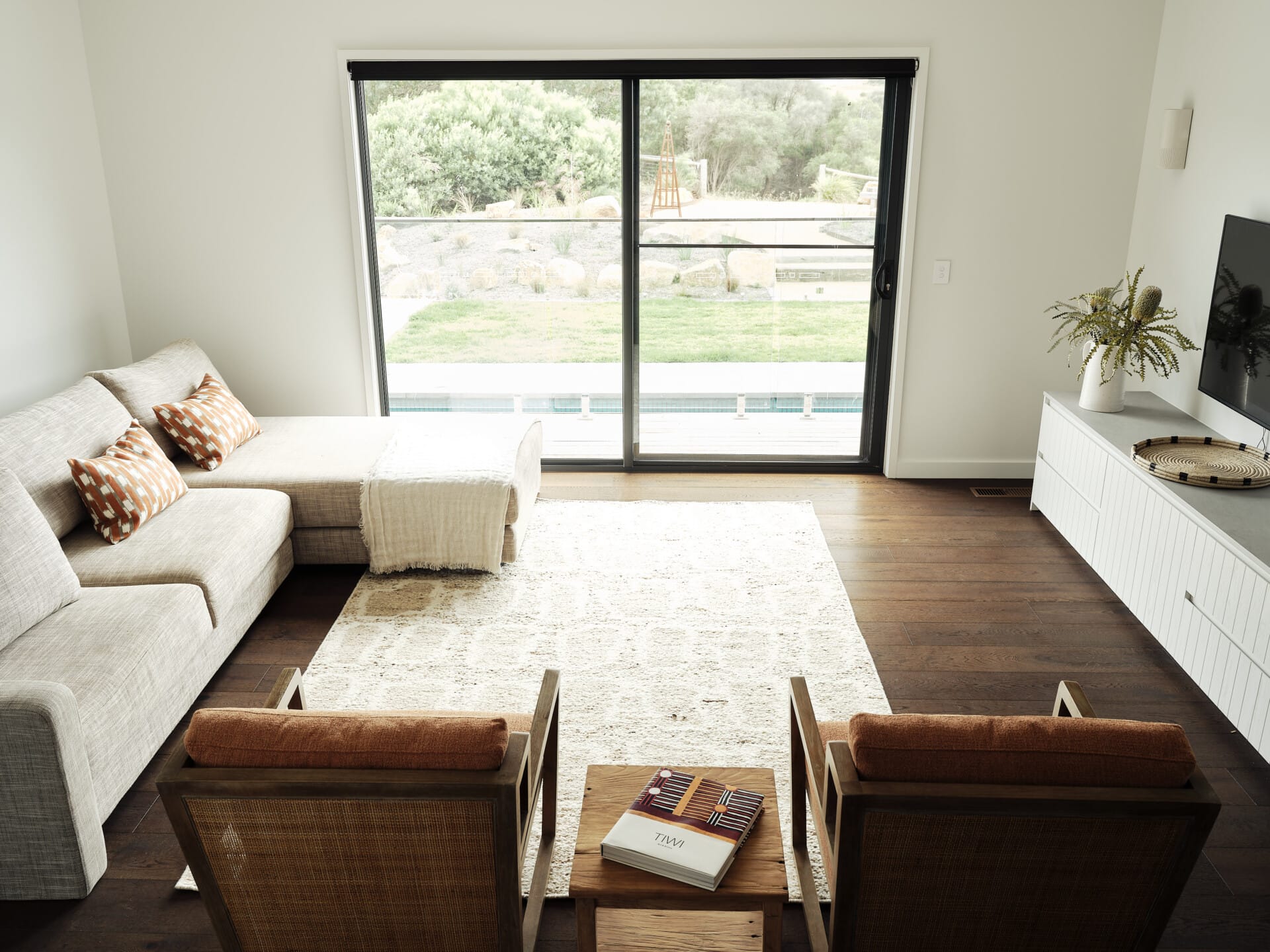 Living room with a sliding glass door overlooking the garden and pool, a linen sofa and rattan armchairs around a timber coffee table, Le Cru 190mm timber flooring underfoot.