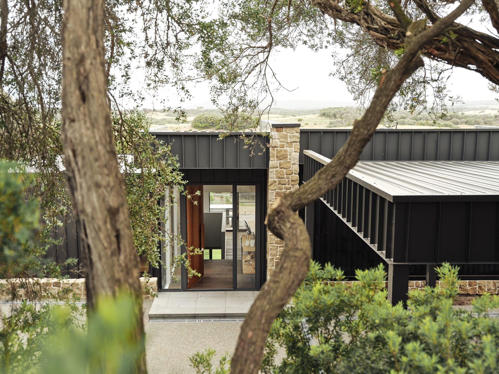 Exterior of St Andrews Residence, with a stone feature column and dark vertical-panelled wings framed by native trees, with an open glass entry and rural views beyond with a glimpse of Le Cru 190mm timber flooring through the glazed entry.