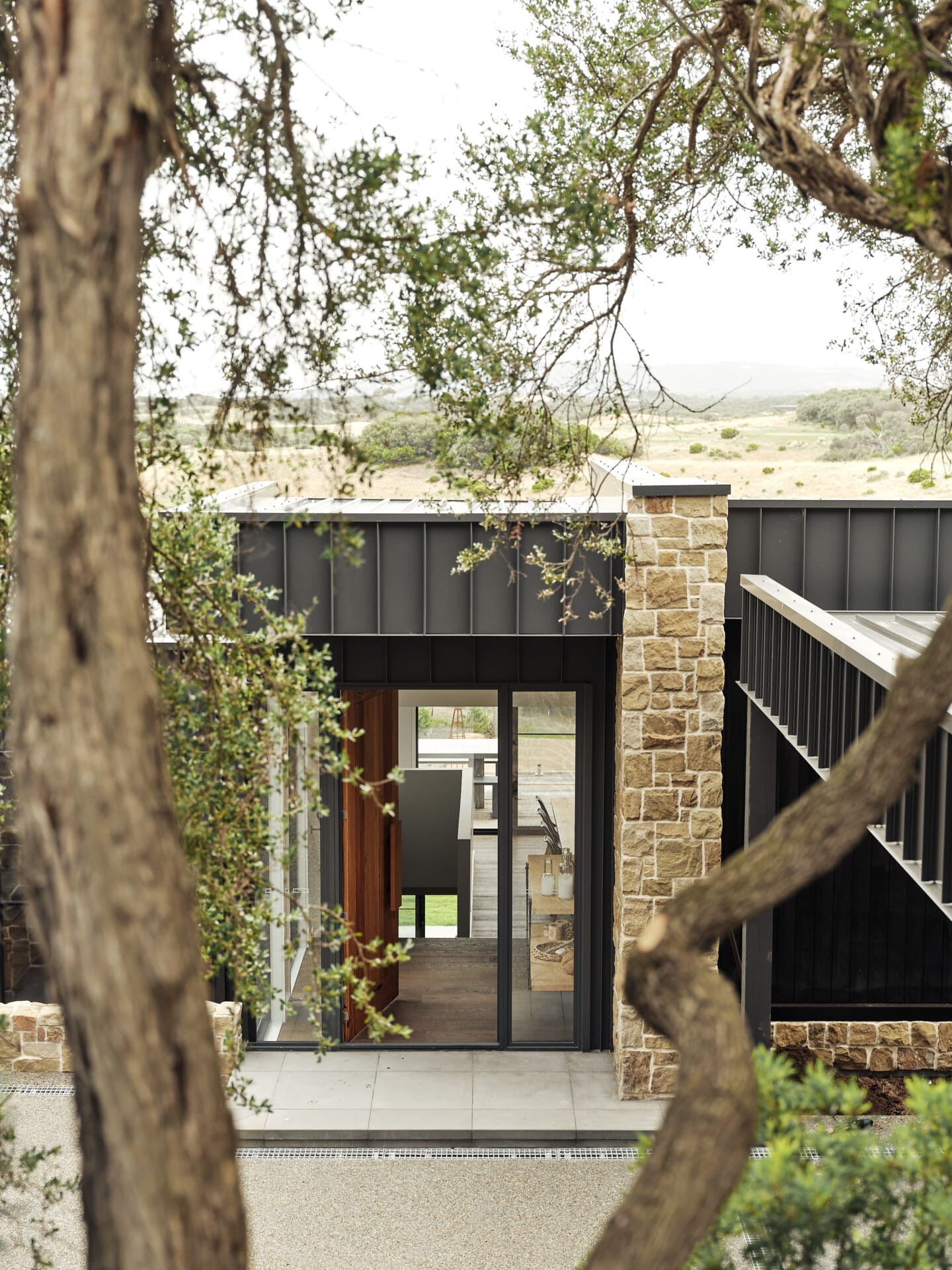 Exterior view through tree branches of the home's stone column and dark cladding, with a sliding door open to the dining area and the golf course landscape beyond.
