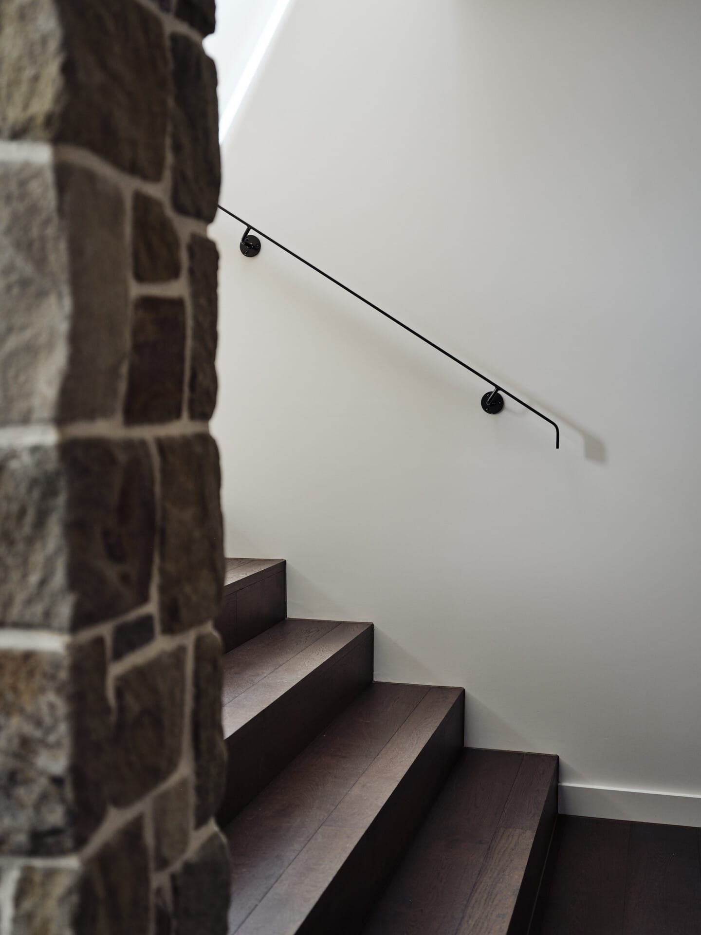 Close-up of a minimalist black handrail against a stone wall, leading down Le Cru 190mm timber stairs.