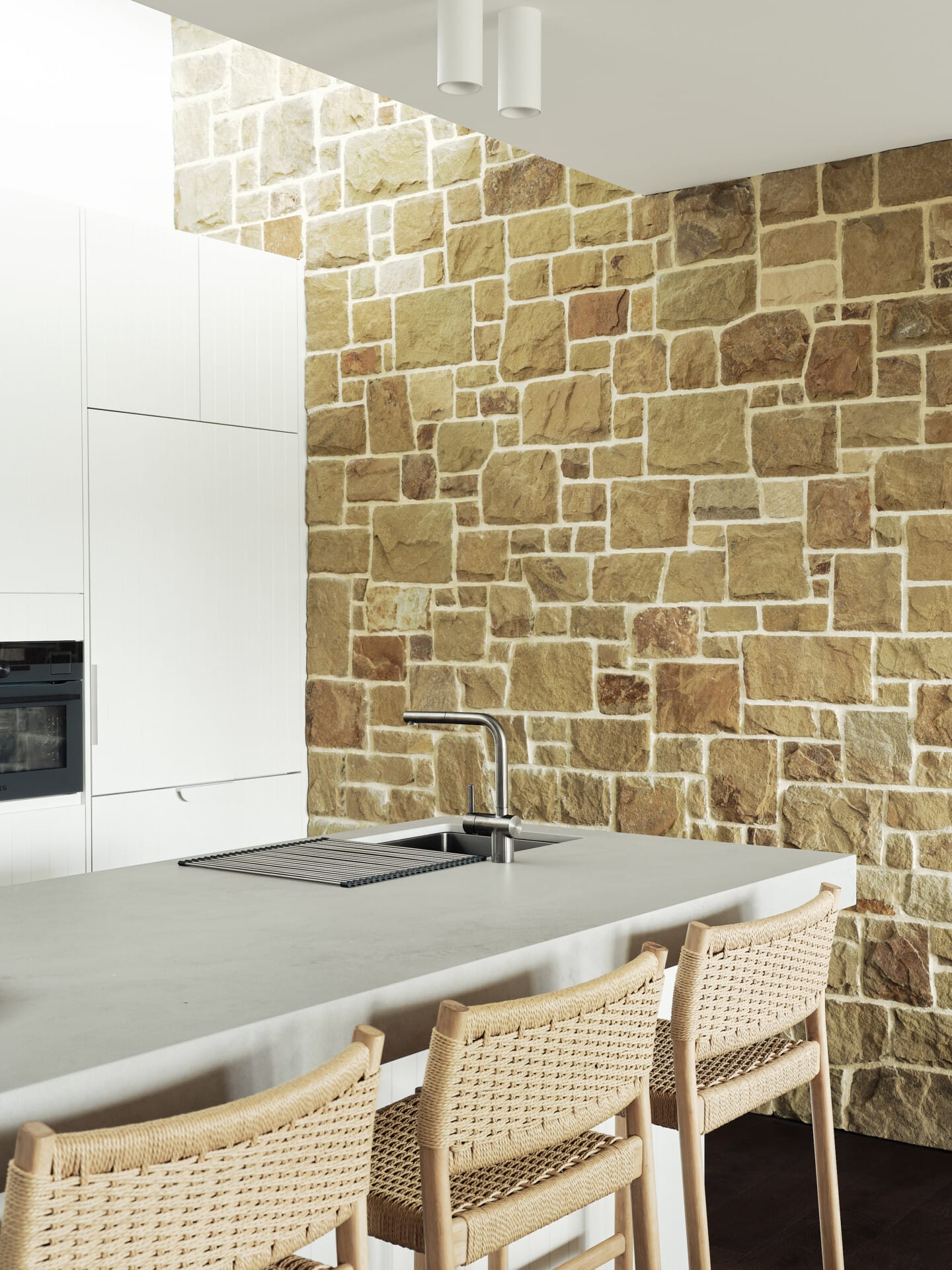 Kitchen island with a stone feature wall, integrated sink and tapware, and woven bar stools along the concrete benchtop.