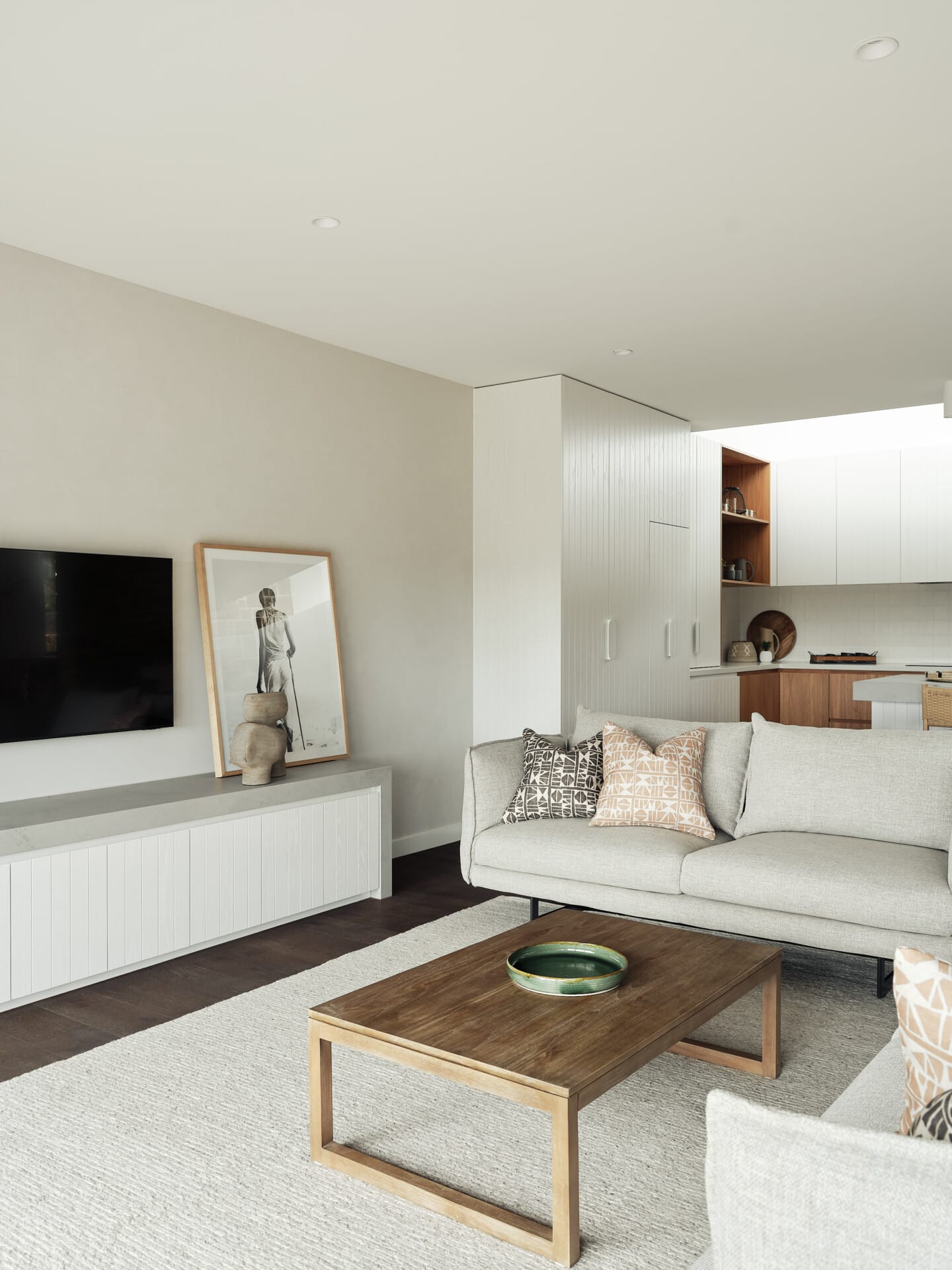 Open-plan living room with a cream sofa and rug, timber coffee table, and framed artwork beside a mounted TV, opening onto the kitchen.
