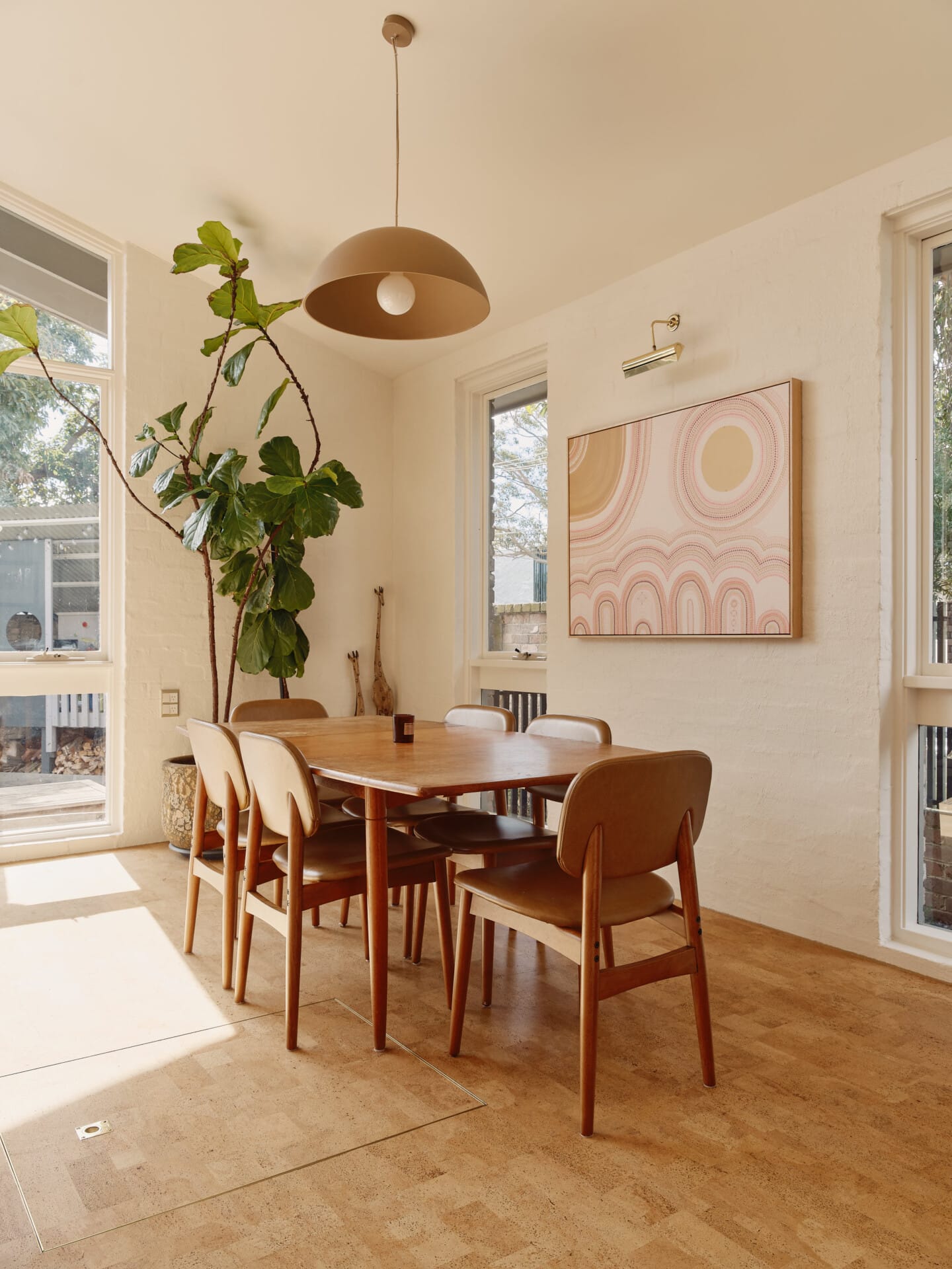 Dining nook with a timber table and mid-century chairs beneath a dome pendant light, a fiddle-leaf fig and framed abstract artwork, Lisbon cork tile flooring throughout.