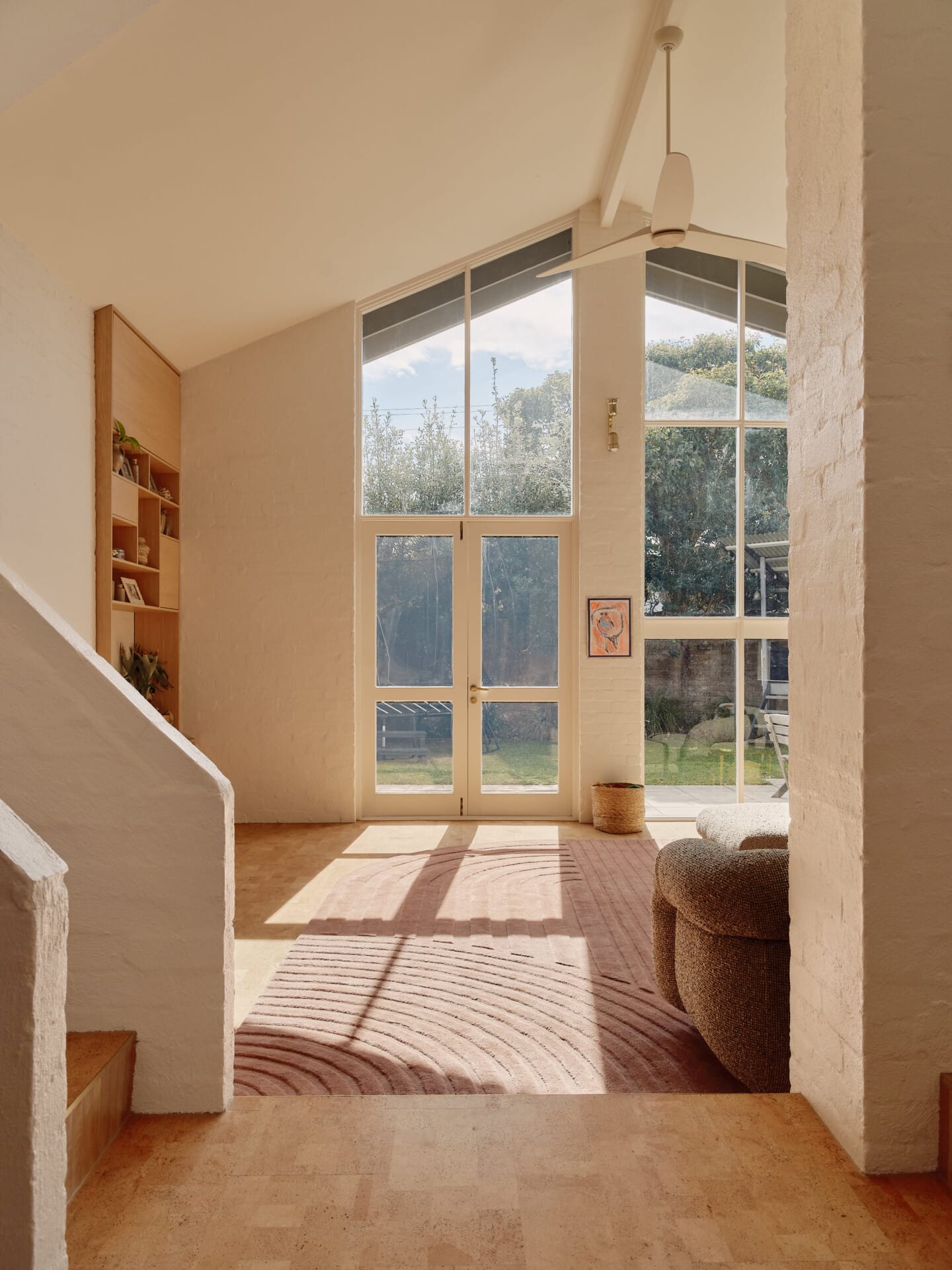 Vaulted living room with French doors opening to the garden, morning light across the Lisbon cork tile floor and a striped rug, a boucle armchair and staircase in the foreground.