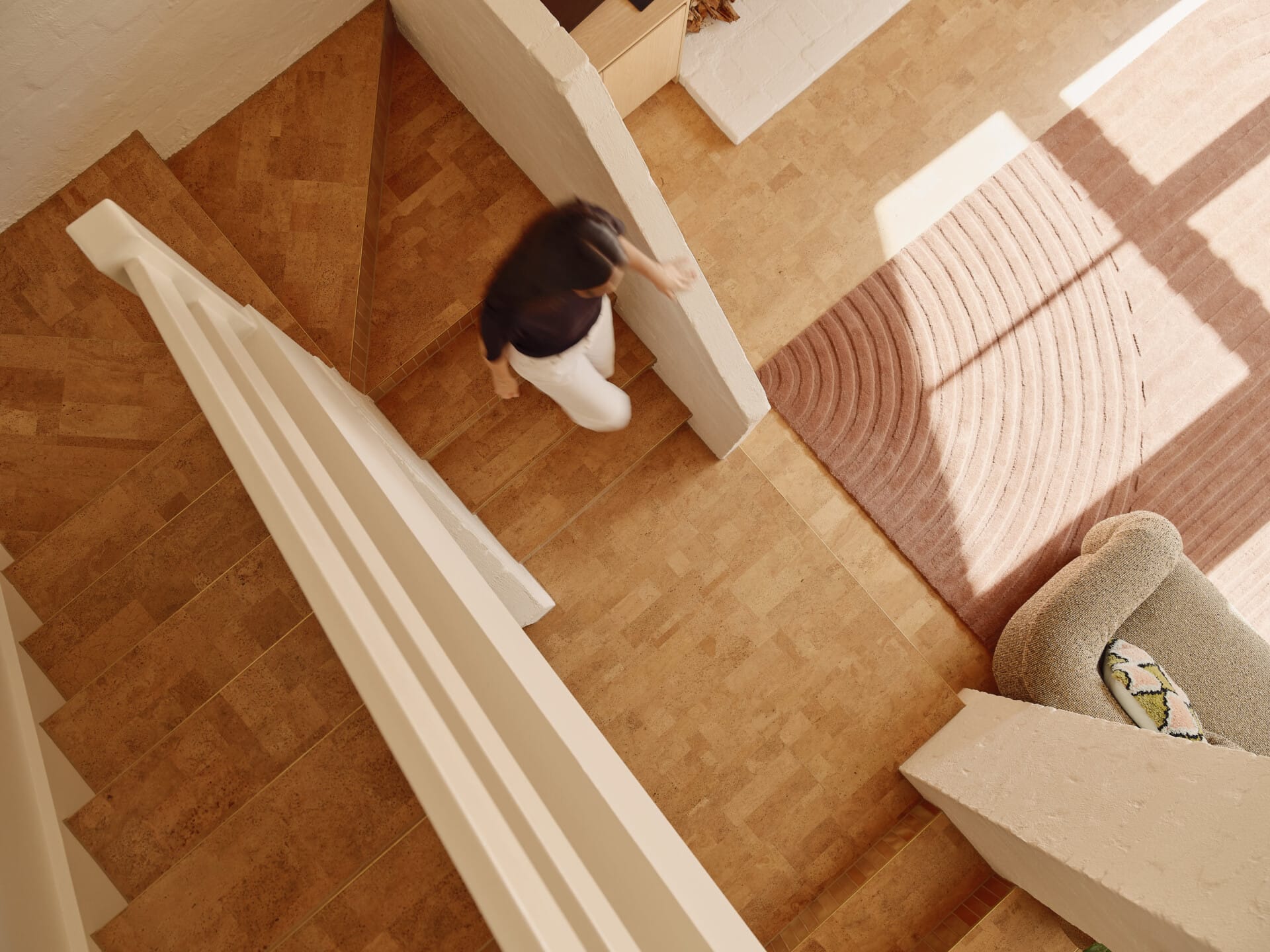 Bird's-eye view down a Lisbon cork staircase and landing, a woman walking toward a sunlit room with a rectangle pink rug and cream armchair.