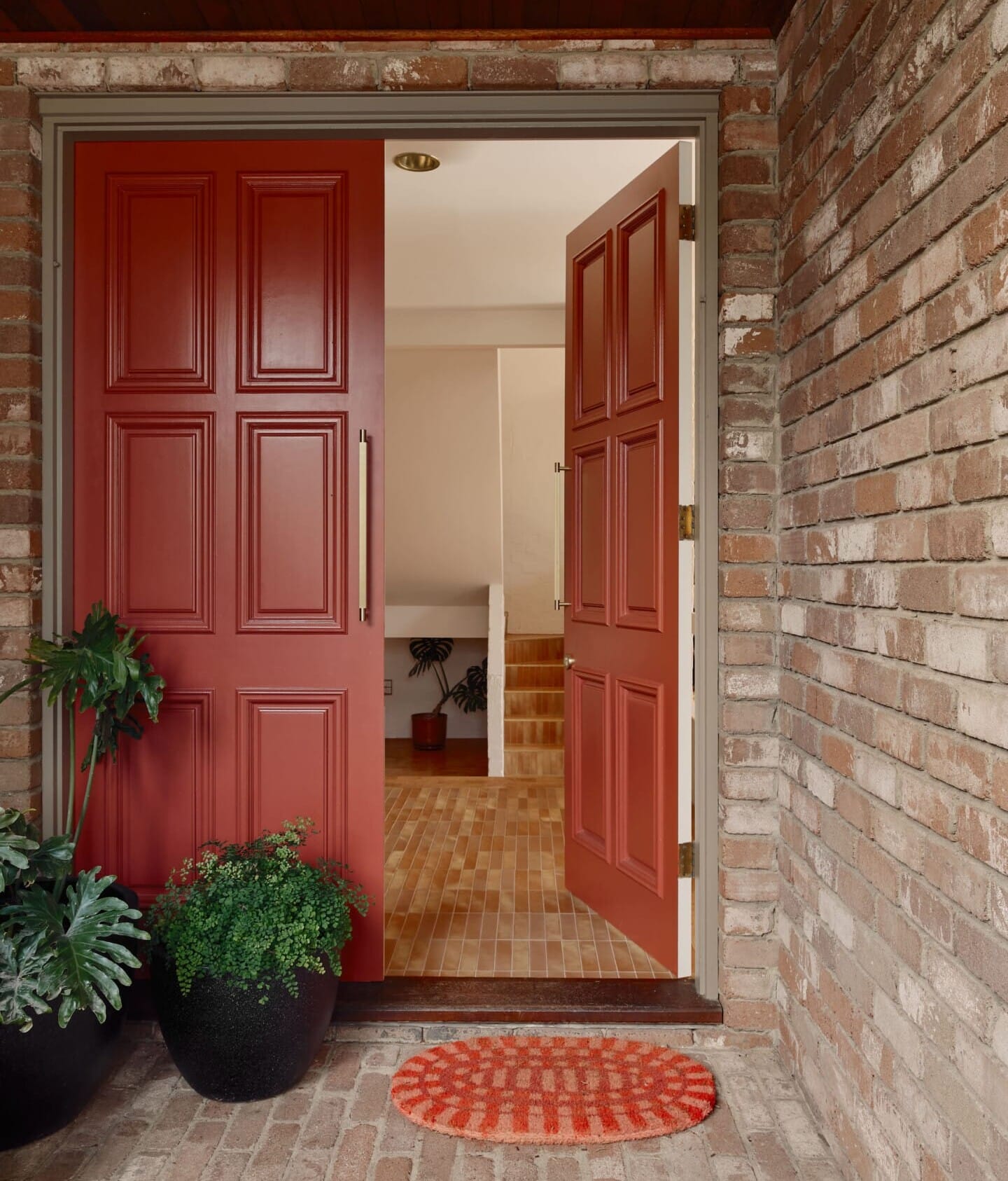Open red front door of a brick exterior, looking through to a hallway floored in Lisbon cork tile, with a staircase and potted plant visible beyond.