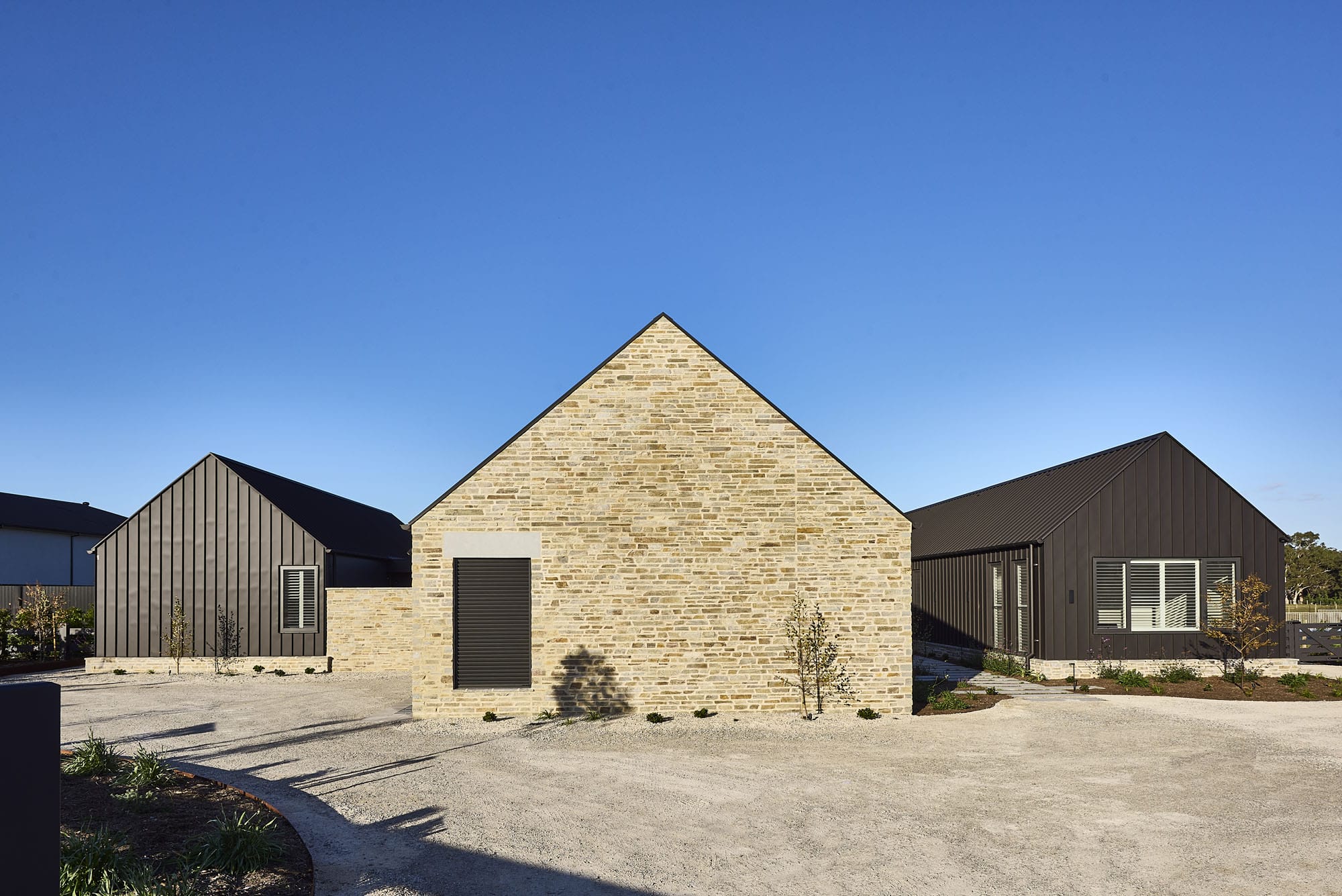 Full exterior view of Riverstone House, showing the stone-clad central gable flanked by dark vertical-panelled wings across a gravel forecourt.
