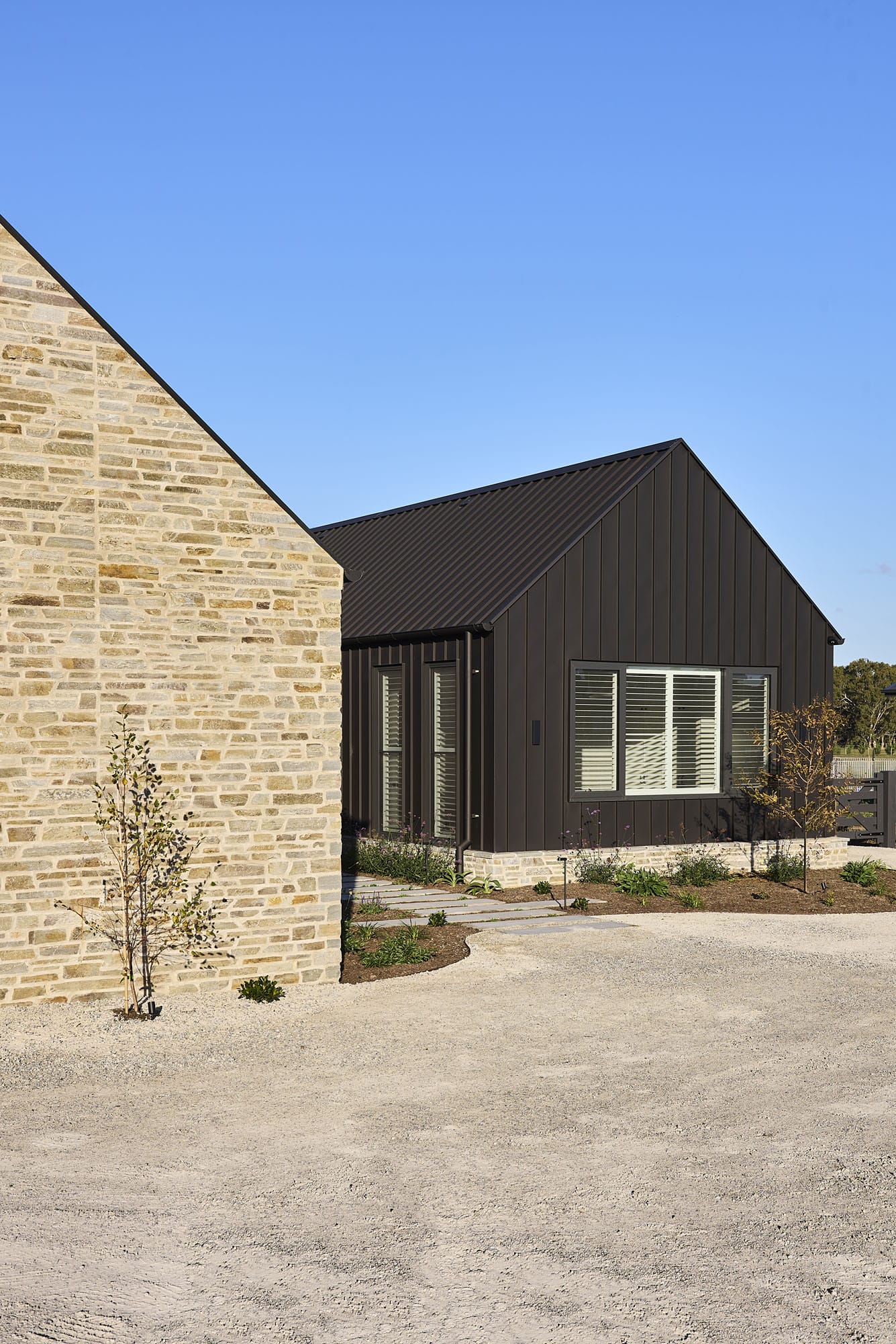 Exterior of Riverstone House, showing the stone-clad wing meeting the dark vertical-panelled gable with plantation shutters, set against a gravel driveway.