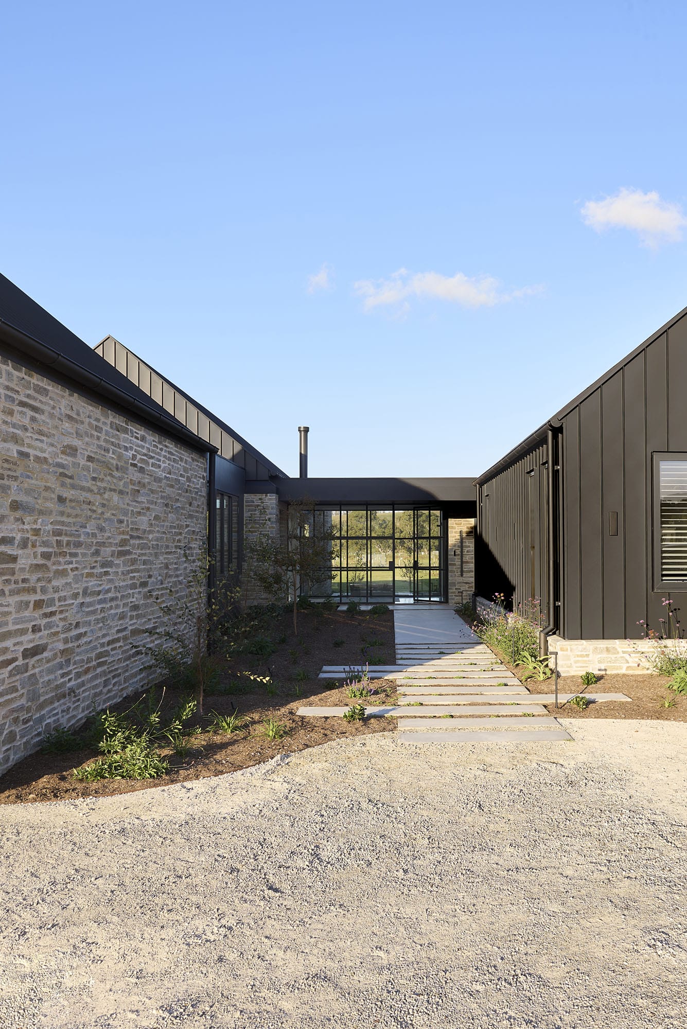 Exterior walkway at Riverstone House, with stepping-stone path leading between the stone-clad and dark vertical-panelled wings to a glazed entry.