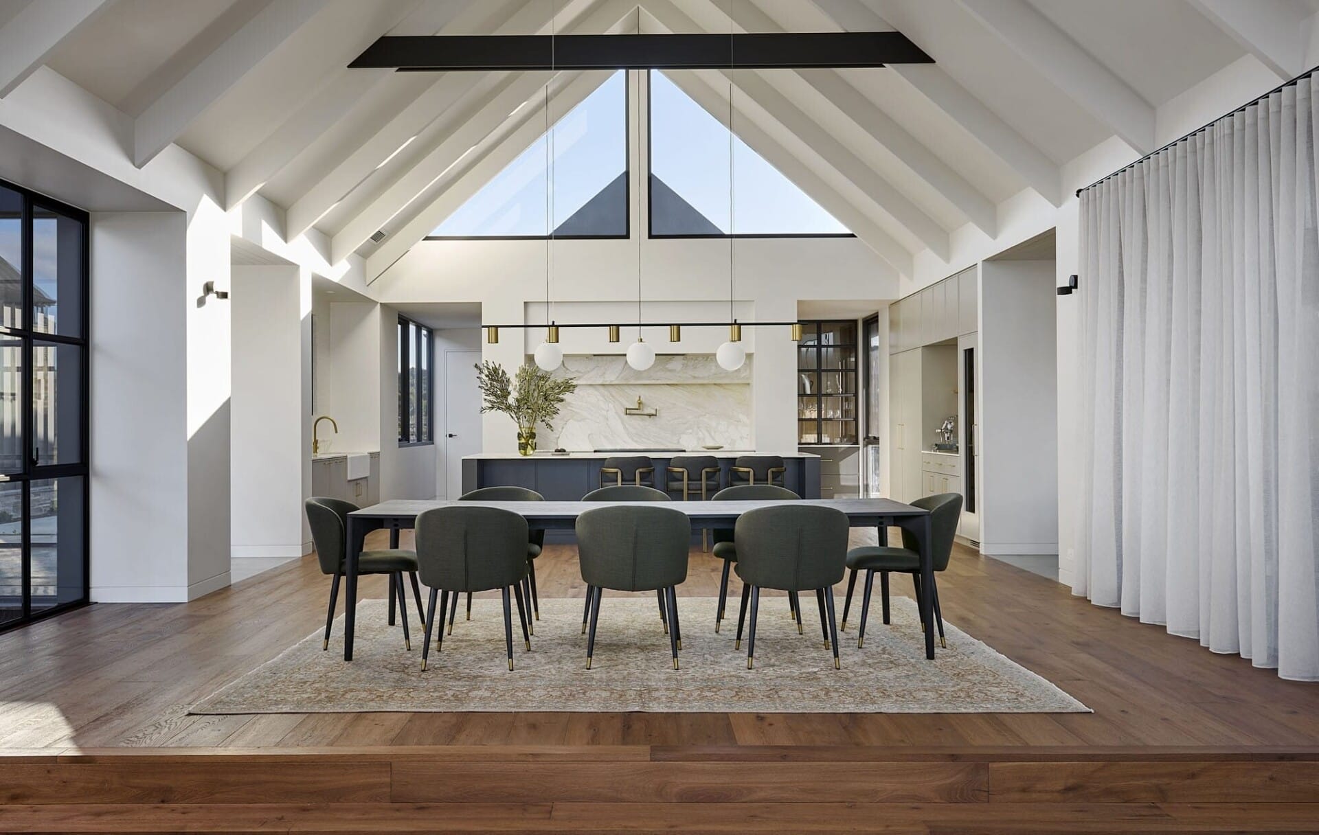 Open-plan dining and kitchen under a vaulted timber-beamed ceiling, with a dark dining suite on a rug and rich Diamond Bay timber flooring extending into a raised entry platform at Riverstone House.