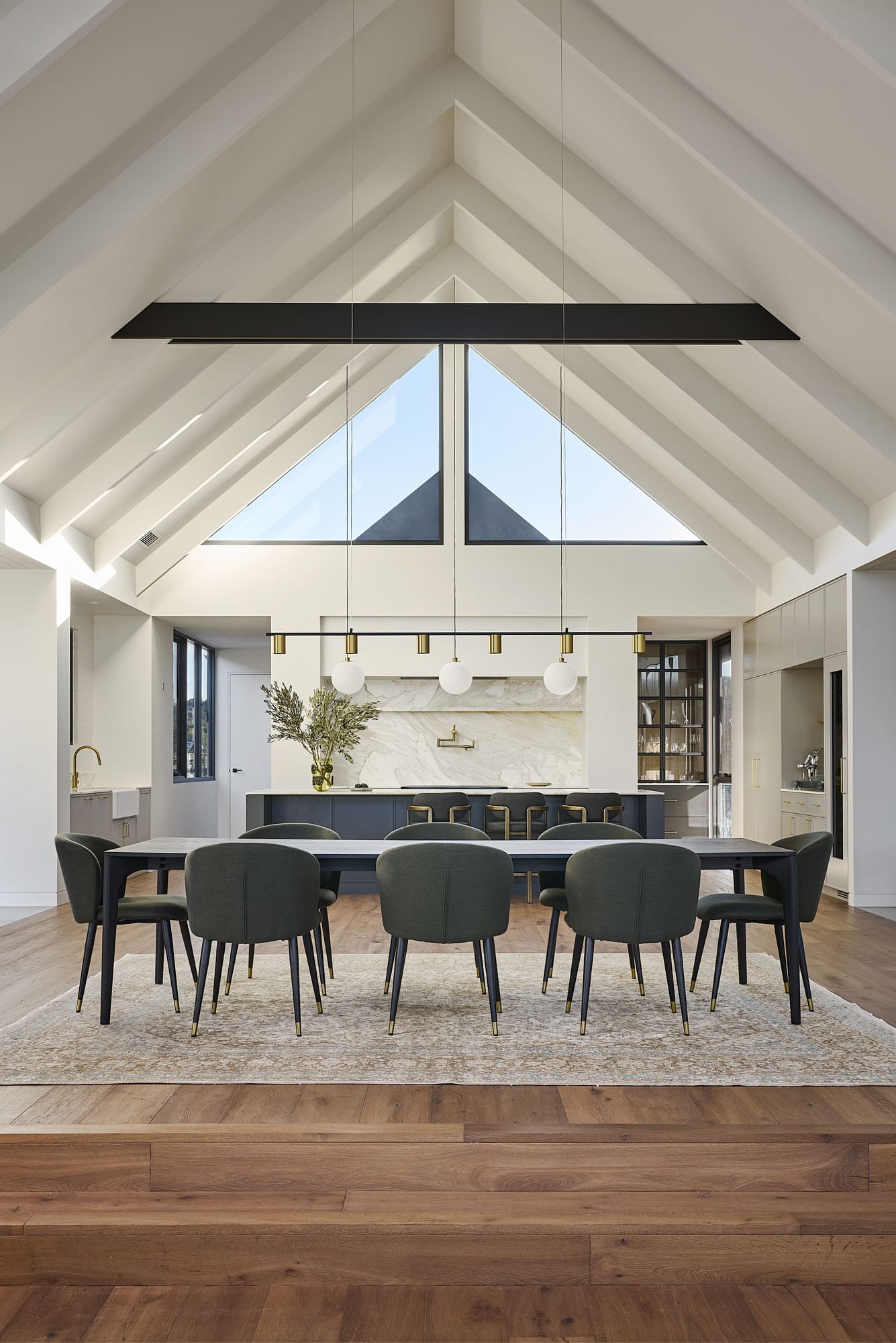 Open-plan dining and kitchen under a vaulted timber-beamed ceiling, with a dark dining suite on a rug and Diamond Bay timber flooring extending into a raised entry platform at Riverstone House.