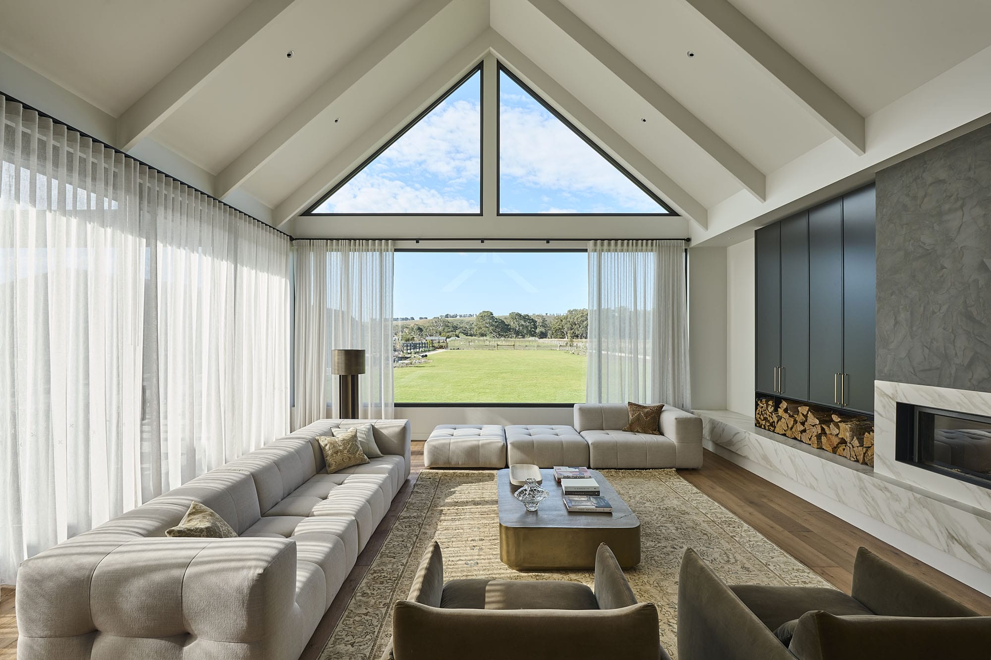 Vaulted living room with a gable window framing paddock views, sheer curtains, a stone fireplace, and Diamond Bay timber flooring at Riverstone House.