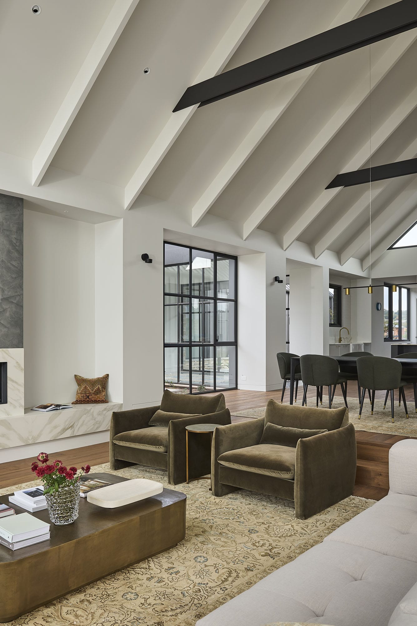 Open-plan living and dining area under a raked timber ceiling, with velvet armchairs, a leather coffee table, and Diamond Bay timber flooring through to the dining space at Riverstone House.