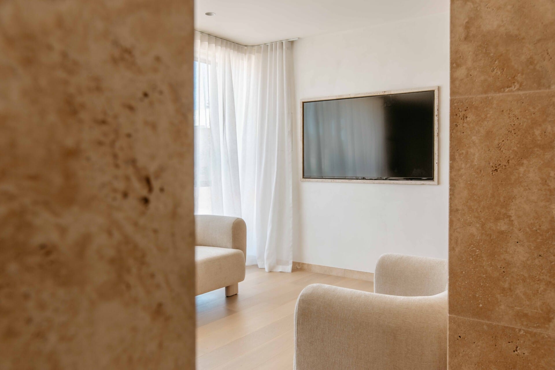 Hotel Sorrento guest suite lounge floored in Kustom Timber Habitat Calypso 190mm engineered oak, viewed through a travertine-clad doorway toward a sheer-curtained window.